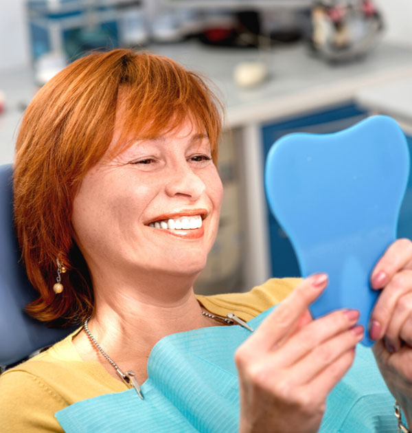 Lächelnde rothaarige Patientin betrachtet ihre Zähne im Handspiegel