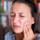 Frau mit Zahnschmerzen greift sich mit der linken Hand an den Kiefer und verzieht dabei das Gesicht.