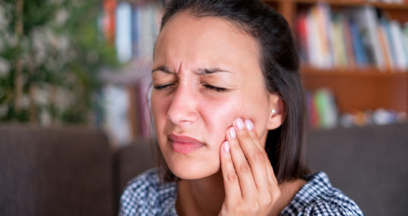 Frau mit Zahnschmerzen greift sich mit der linken Hand an den Kiefer und verzieht dabei das Gesicht.